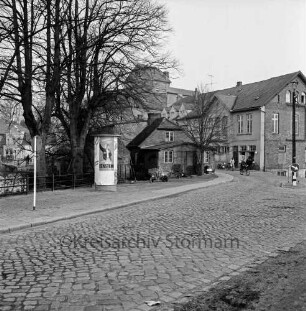 Matthias-Claudius-Straße / rechts Kreuzung mit Klosterstraße: Kopfsteinpflaster: Bildmitte Kocksche Schmiede, Baum, Fahrradfahrer: im Hintergrund Claudius-Mühle: links Seepromenade, Litfaßsäule, Geländer, Bäume, dahinter Herrenteich: rechts Verkehrsinsel mit Straßenlampe, Verkehrsschild, Wegweiser