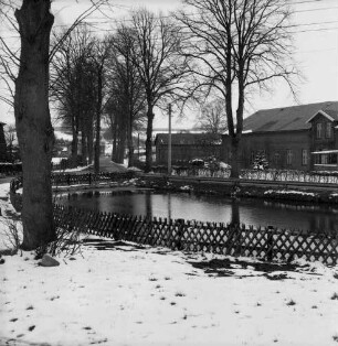 Dorfteich mit Jägerzaun: dahinter Bauerngehöfte und Bäume: Winter