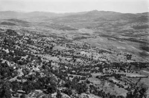 Dorf und Landschaftsblick : Blick über Getreidefelder unter Öl- und Feigebäumen sowie Eichen, Eschen und Macchiebüschen in der Nähe eines kabylischen Dorfes, das in einem Tal im Département de Tizi Ouzou gelegen ist.
