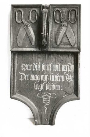Sammelbrett der Lübecker Schneiderzunft. Das Brett diente zum Sammeln der Umlagen und Strafgelder. Holz, geschnitzt, Scherenrelief, Holzgriff, Spruch-Ornament