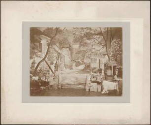 Bauernhofkulisse, davor Stand mit Keramik, Kinderspielzeug, Körben und hölzernen Gerätschaften.
