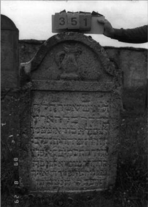 Grabstein 351 (Index: 100107798) Material: Kalkstein Zustand: verwittert Maße HxBxT: 127x68x18 cm Beschreibung: Stele mit geschwungen eingezogenem Rundbogenabschluß, Schriftfeld rechteckig eingetieft Symbolik: Levitenkanne und -schale: Grabstein 351