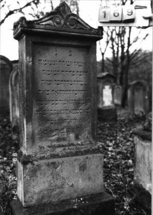 Grabstein 169 (Index: 99752009) Material: Sandstein Zustand: verwittert Maße HxBxT: 150x50x21 cm Beschreibung: Stele, Sock. , Abdeckplatte m. Volutenaufsatz m. Palmette, rechteck. Schriftfeld einget Symbolik: Voluten, Palmette, Blätter: Grabstein 169