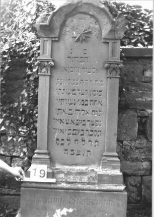 Grabstein 19 (Index: 100114428) Material: Sandstein Zustand: teilweise abgebrochen, leicht verwittert Maße HxBxT: 167x72x41 cm Beschreibung: Stele, getreppter Sockel, eingezogener Rundbogenabschluß mit Verdachung, beidseitig Pilaster, beschädigte Eckakrotere, Aufsatz fehlt Symbolik: beschädigter Stern, geschmückte Kapitelle: Grabstein 19