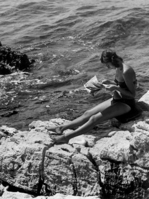 Urlaub in Jugoslawien. Eine Frau liest am felsigen Strand die Tagespresse