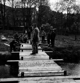 Sühlen: Trave: Wanderweg zwischen Sühlen und Bebensee (Kreis Segeberg): Fußgängerbrücke: Bau: Technisches Hilfswerk (THW) Bad Segeberg: Verlegung der Holzbohlen auf Trägern durch freiwillige Helfer: hinten zwischen Bäumen "Zettlersche Mühle" [ehemalige Kupfermühle] mit Nudelfabrik