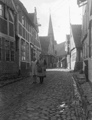 kleine Gasse mit Kopfsteinpflaster, Fachwerkhäuser, Kirchturm und zwei Frauen im Vordergrund