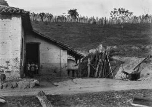 Arbeiterhaus : Ein Arbeiterhaus in Amparo mit einem Backofen unter einem kleinen Dachvorsprung in der Bildmitte, einer Trinkwasserversorgung rechts und drei Kindern am Hauseingang stehend.