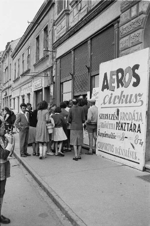 Straßenbild einer Kleinstadt mit Einheimischen an einem Kassenschalter, davor Werbeaufsteller des Zirkus Aeros
