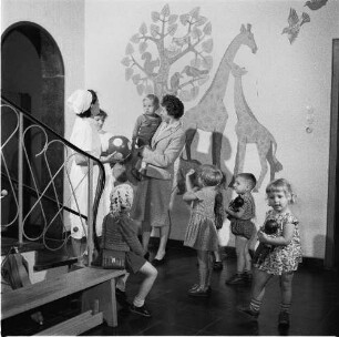 Erzieherinnen in einer Kinderkrippe (oder einem Kindergarten) mit Kindern und einer Mutter, die ihren kleinen Sohn auf dem Arm trägt