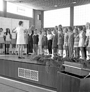 Volksschule: Sporthalle: Neubau: Einweihung: Innenansicht: Blick auf die Bühne: darauf Lehrerin mit Schulchor