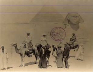 Gizeh, Ägypten. Touristengruppe mit Kamelen und Fremdenführern vor Sphinx und Pyramide von Gizeh