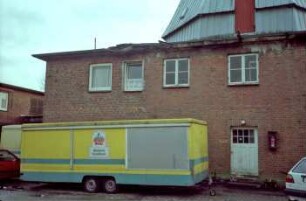 Braaker Mühle: Teilansicht: Unterbau mit Eingangstür, oben Mühlenturm [angeschnitten] mit Tür: davor Wagenanhänger mit Firmenschild "Braaker Mühle - Bäckerei Konditorei"