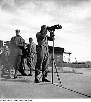 Rostock-Warnemünde. Deutsche Verkehrsfliegerschule, Zweigstelle Seefliegerschule Warnemünde. Ein Pilot beobachtet auf dem Fluggelände durch ein Fernglas das Fluggeschehen (im Hintergrund Gebäude der Ernst Heinkel Flugzeugwerke)
