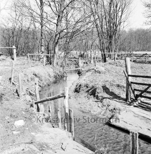 Gölmbach: rechts zwei Gatter: an Uferböschung Holzzaun: hinten Steg: dahinter Bäume: hinten Wald