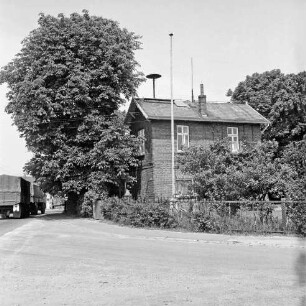 Dorfring: Wohnhaus mit Sirene auf dem Dach: ehemalige Volksschule: davor Vorgarten mit Hecke und Zaun: links davor Kastanienbäume: links Landesstraße Richtung Autobahn A 1 und Stapelfel mit Lkws