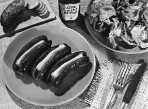 Krafts Rezeptaufnahmen. Mit Käse gefüllte Bratwürste. Daneben ein Brettchen mit aufgeschnittener saurer Gurke, eine Flasche Kraft's Tomatenketchup und eine Schüssel Salat