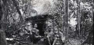 Zwei Frauen, vermutlich Gyele, mit zwei Kindern vor einer Hütte im Urwald