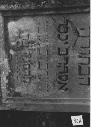 Grabstein 92 (Index: 100106472) Material: Sandstein, Marmorrosette Zustand: verwittert und abgeblättert, teilweise Textverlust, Maßwerk nachgemalt, Mittelaufsatz fehlt, Lettern falsch nachgezogen Maße HxBxT: 131x51x15 cm Beschreibung: Stele mit Walmdach und Mittelaufsatz, Mittelpalmette, Schriftfeld eingetieft in Maßwerkform, getreppter Sockel: Grabstein 92A