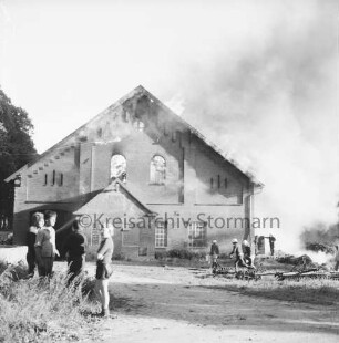 Gut Altfresenburg: Brand des Bullenstalls: Brandruine: Giebelfront: rechts Eggen: dahinter Feuerwehrleute beim Verlegen des Wasserschlauchs und weitere Personen, Rauchschwaden: links vier Kinder: dahinter Bäume: 24. August 1971