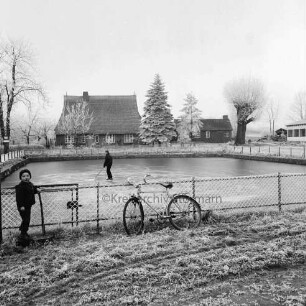 Dorfteich: im Vordergrund Kind an Zaun, daneben Fahrrad: Schlittschuhläufer auf Teich: im Hintergrund Bäume und Häuser: Winter