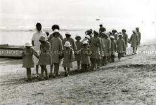 Kindergruppe am Strand, alle mit karierten Kleidern und weißen Hüten bekleidet