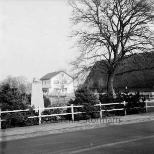 Dorfring: Ehrenmal 1939-1945 mit Grünanlage: davor Zaun: rechts Friedenseiche: dahinter Bauernhaus mit Reetdach: hinten links Zweigstelle der Kreissparkasse Stormarn