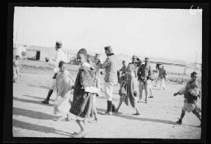 "Maurenstadt" neben dem Lager von Nador (Marokko): Einheimische Kinder und Offiziere [des spanischen 8. Kavallerie-Regiments "Lusitania"] in hellgestreiften Uniformhosen, dunklen Uniformjacken und niedrigen, runden Feldmützen auf einem Platz mit Gebäuden im Hintergrund [Eröffnung der Bahnstrecke Nador-Zeluan (Selouane, Marokko]