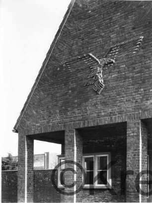 Adlerrelief: fliegender Adler, in Fängen Hakenkreuz: an Giebel von Backsteingebäude: darunter Arkaden: im Hintergrund links Gebäude;