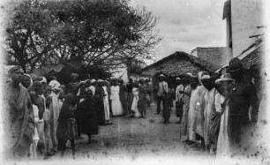 In Bagamoyo : Straßenszene in Bagamoyo. Schaulustige haben sich auf der Straße versammelt, in der Bildmitte steht einer der Forschungsreisenden mit Schirm und Tropenhelm