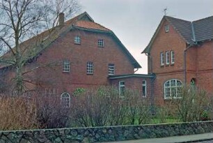 Hauptstraße 64: Hofanlage: rechts Wohnhaus aus Backstein [angeschnitten] mit Quergiebel, Sprossenfenster: links giebelständiges Wirtschaftsgebäude, Giebelseite mit Sprossenfenstern: Bildmitte flacher Verbindungsbau: davor Garten, im Vordergrund Hecke, Feldsteinmauer