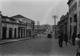 Straßenszene in Catanduva : Straße von Bahnhof in die Stadt Catanduva hinein mit Gebäude und Bürgersteigen. Einige Passanten sind unterwegs, im Mittelgrund Fahrzeuge auf der Straße. Links ein Hotel, rechts ein Werbeschild für den Mineralölkonzern "Esso".