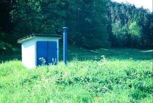 modernes Wasserwerk mit blauer Messsäule davor auf einer Wiese