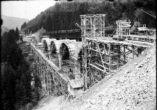 Alter Viadukt mit einem darauf befindlichen Eisenbahnzug, Gerüst mit Aufzug zum Ablassen von Steinen und im Bau befindliche Bögen