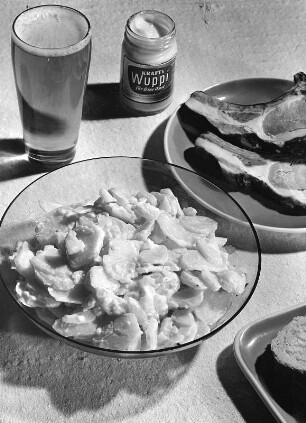 Krafts Rezeptaufnahmen. Schale mit Kartoffelsalat, Teller mit Koteletts, Brotteller, Kraft's Wuppi und Bierglas