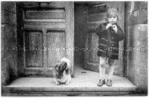 Kleines Mädchen steht auf einer Türschwelle, hinter sich die halboffene Eingangstür, neben sich ein Pekinese