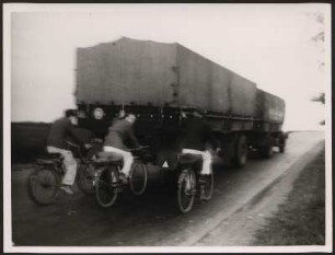 Drei Fahrradfahrer fahren hinter einem LKW her, anscheinend hält sich die mittlere Person am Anhänger fest, was sehr gefährlich für den Verkehr sein kann