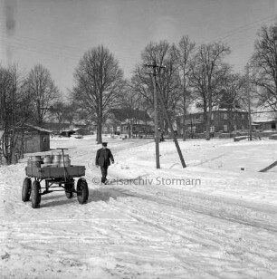 Dorfstraße mit Fußgänger: links Anhänger mit Milchkannen: im Hintergrund Bauernhäuser und Bäume: Winter