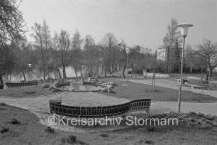 Am Jungfernstieg: Grünanlage mit kleinem Teich, Bänken, Lampen und Wegen: hinter Baumreihe Neuhöfer Teich: dahinter rechts Häuser
