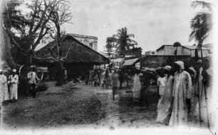 In Bagamoyo : Straßenszene in Bagamoyo. Blick über einen Platz mit vielen Schaulustigen. Am linken Bildrand steht einer der Forschungsreisenden mit Schirm und Speer