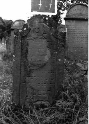 Grabstein 77 (Index: 100103575) Material: Sandstein Zustand: stark verwittert, abgeblättert, weitgehender Textverlust Maße HxBxT: 100x48x8 cm Beschreibung: Stele, eingezogener Rundbogenabschluss, Schriftfeld eingetieft mit Konvexbogenabschluss Besonderheit: ergänzt nach Gräberbuch: Grabstein 77
