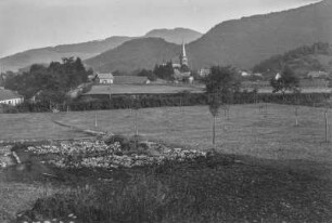 Dorf : Blick auf ein Dorf in einer Hochebene. Im Hintergrund bewaldete Berghänge und eine Kirche, im Vordergrund Felder und Weiden