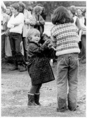 Kind in Gummistiefeln und Regenmantel neben einer Matschpfütze, es zeigt einem älteren Kind etwas auf der Hand, im Hintergrund Menschen (Altersgruppe 18-21)
