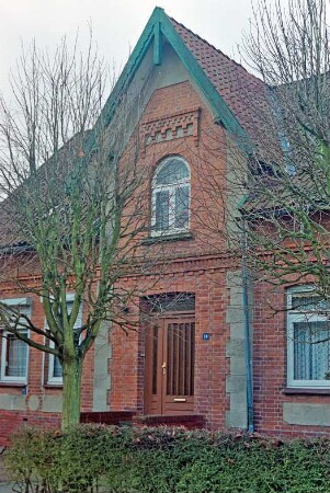 Sattenfelde: Bahnhofstraße 38: traufständiges Backsteinhaus: Straßenfront, Teilansicht: Mittelrisalit mit Dreiecksgiebel, Eingangstür, Hausnummer: davor Bäume, Hecke