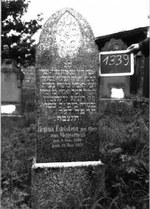 Grabstein 1339 (Index: 100104638) Material: Granit Zustand: gut erhalten Maße HxBxT: 110x45x18 cm Beschreibung: aufgesockelte Stele mit Spitzbogenabschluss, seitlich eingezogen, im Bogenfeld Ornamente (stilisierte Blüten?) Symbolik: stilisierte Blüten?: Grabstein 1339