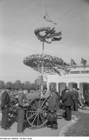 Besucher an einem landwirtschaftlichem Gerät sowie Richtkranz auf der Technischen Messe 1953