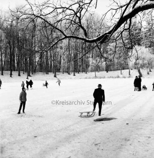 Kurpark: Salzteich: vorn Mann mit Schlitten: hinten rechts Paar mit Schlitten: links mehrere Kinder mit Schlittschuhen und Schlitten auf Eisfläche: hinten Bäume: Winter