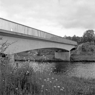 Trave: vorn Böschung mit Blumen: dahinter Brücke der Landstraße Richtung Reecke (Hansestadt Lübeck): am gegenüberliegenden Ufer rechts an Böschung Treppe: dahinter zwischen Bäumen der Krichturm