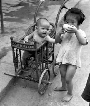 Schwester, aus einer Schale trinkend, und Bruder, in einem Bambus-Kinderwagen sitzend, am Straßenrand