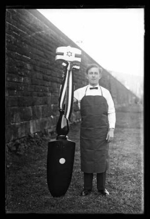 Mann mit Schürze, in der Hand eine Art Standarte (oben mit sechseckigem Stern) mit Schleife geschmückt
